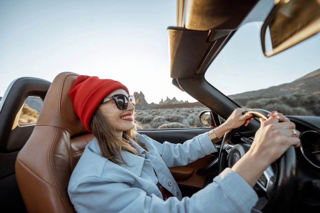 A smiling woman wearing a red beanie and sunglasses drives a convertible through a scenic desert landscape under a clear sky.