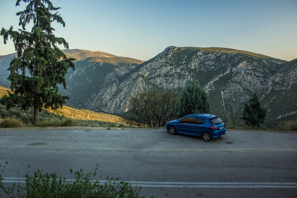 A blue car is parked on the side of a mountain road with tall trees and rocky hills in the background.