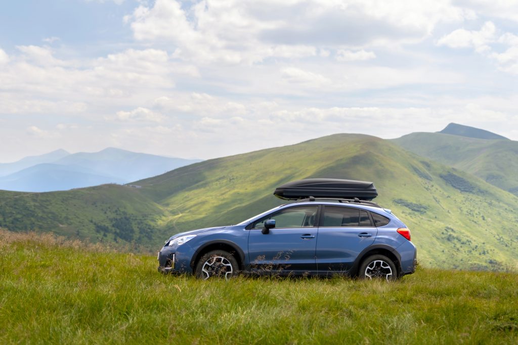 A blue SUV with a rooftop cargo box is parked on a grassy hilltop, surrounded by scenic, rolling green mountains under a partly cloudy sky.