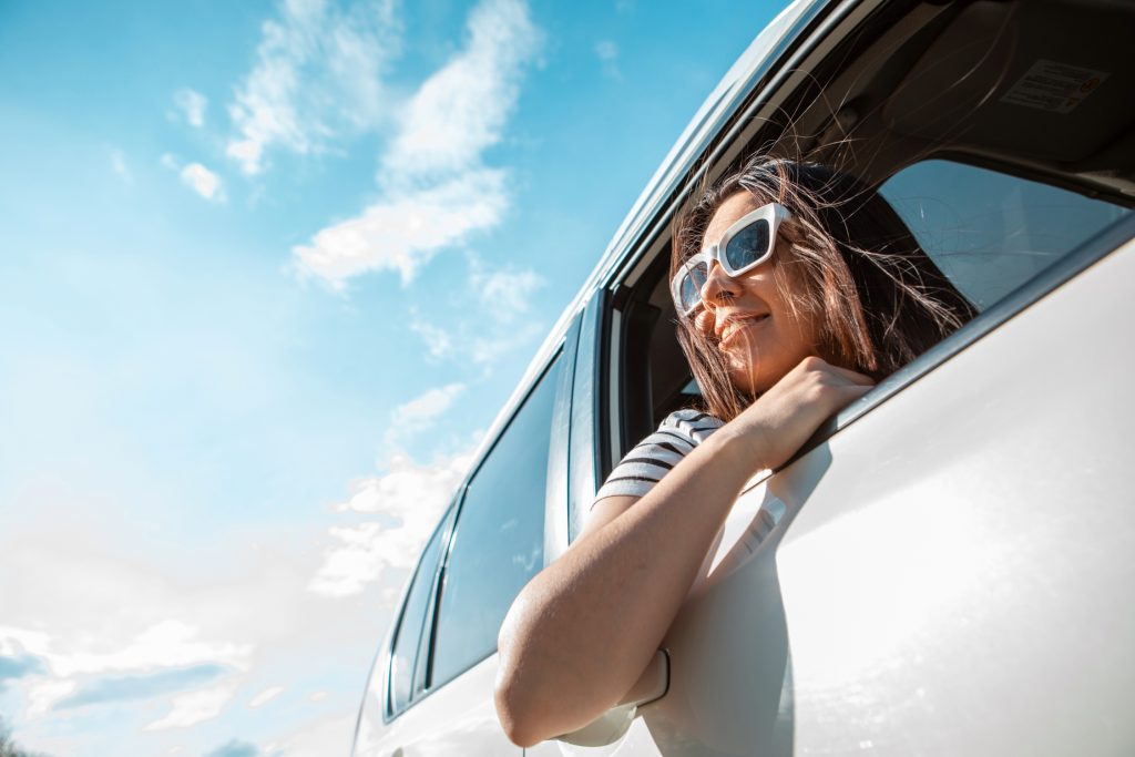 A young woman leans out of a white car window smiling under a bright blue sky.