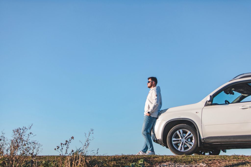 A man in casual attire leans against a white SUV parked on a grassy hill, gazing into the distance under a clear blue sky.