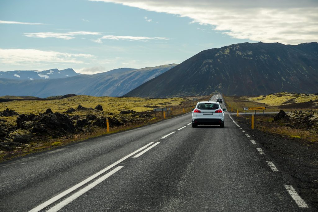 A car drives along a scenic highway surrounded by mountains and rugged volcanic terrain.