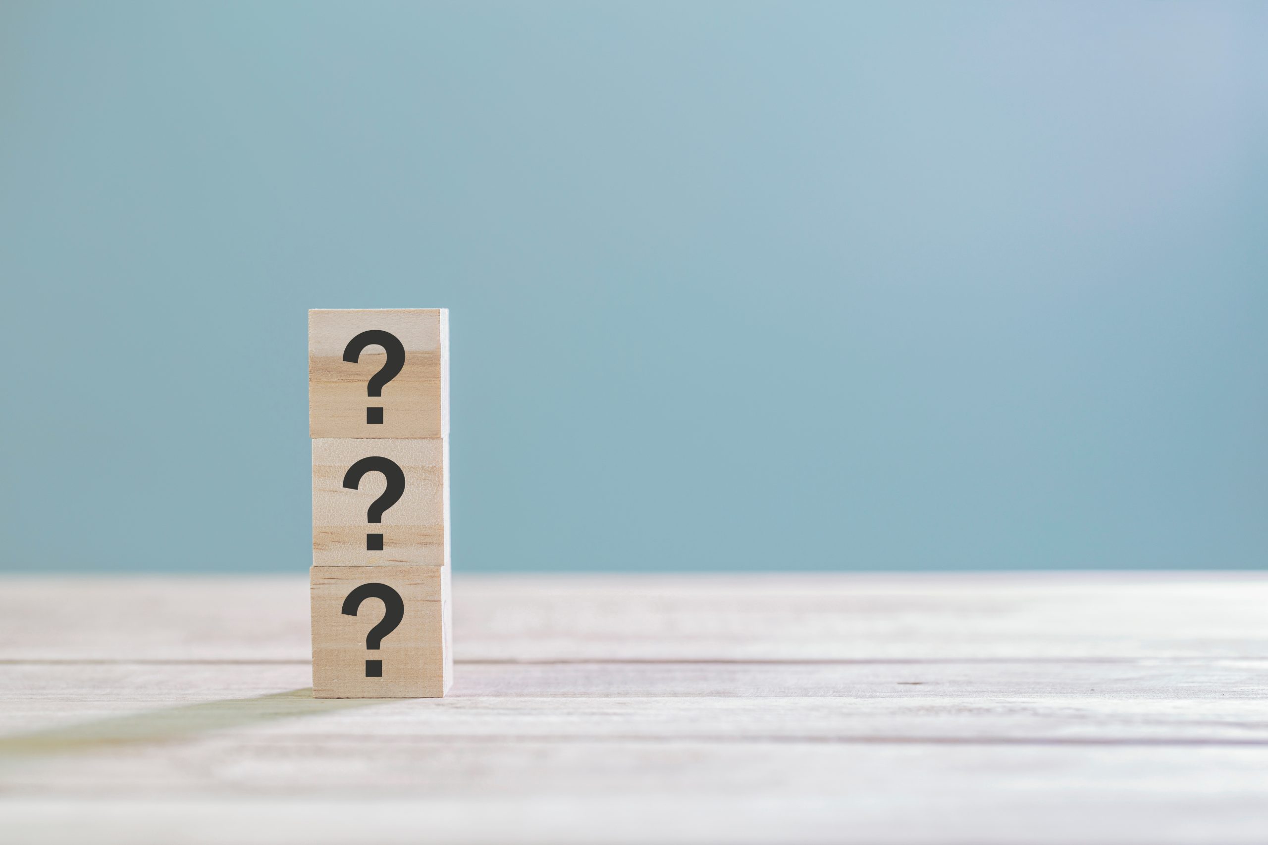 Three wooden blocks stacked vertically, each featuring a black question mark, placed on a light wooden surface against a soft blue background. The image symbolizes curiosity, uncertainty, or frequently asked questions.