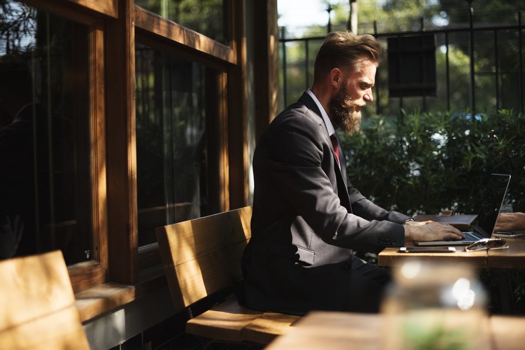 well-dressed man in a dark suit working on a laptop at an outdoor wooden table.