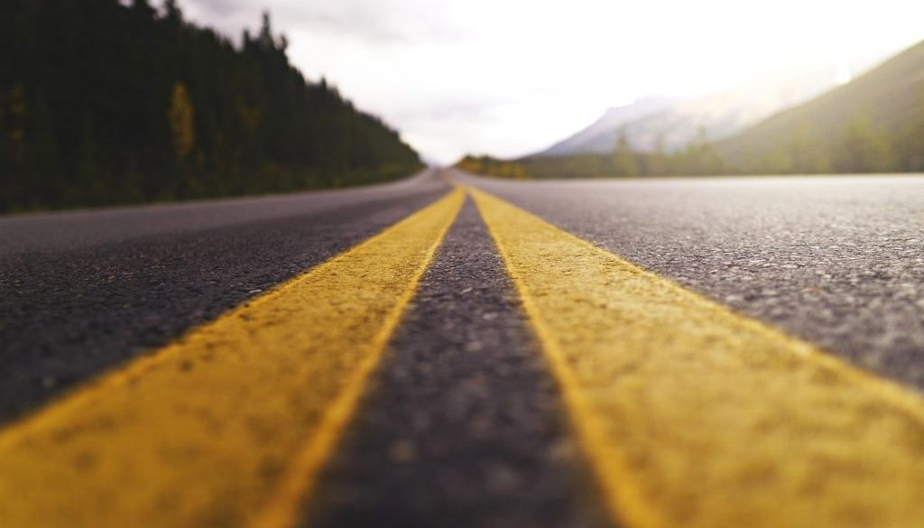 A low-angle view of a Montana highway captures the bold yellow centerlines stretching toward distant mountains, framed by dense pine forests and glowing in the golden light of dusk or dawn.