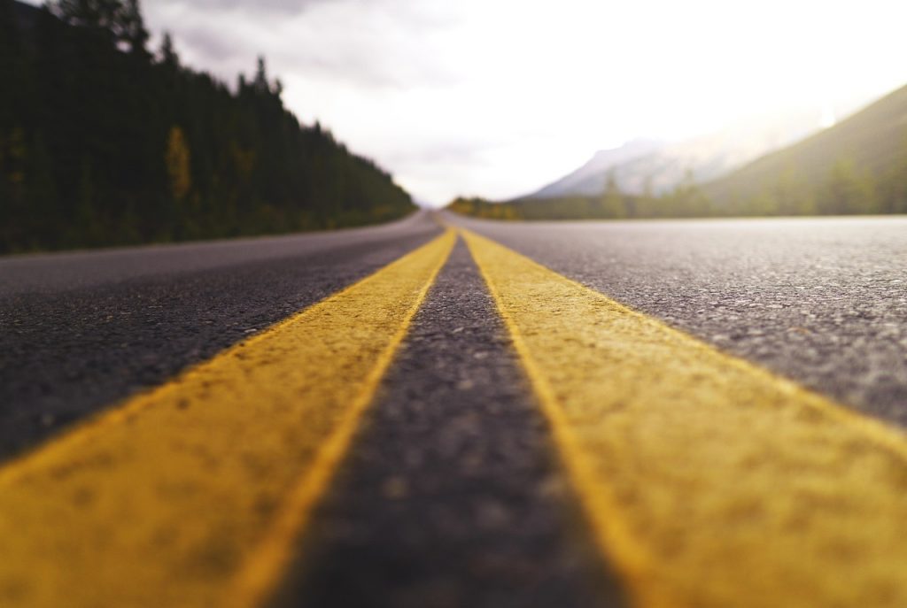 A low-angle view of a Montana highway captures the bold yellow centerlines stretching toward distant mountains, framed by dense pine forests and glowing in the golden light of dusk or dawn.
