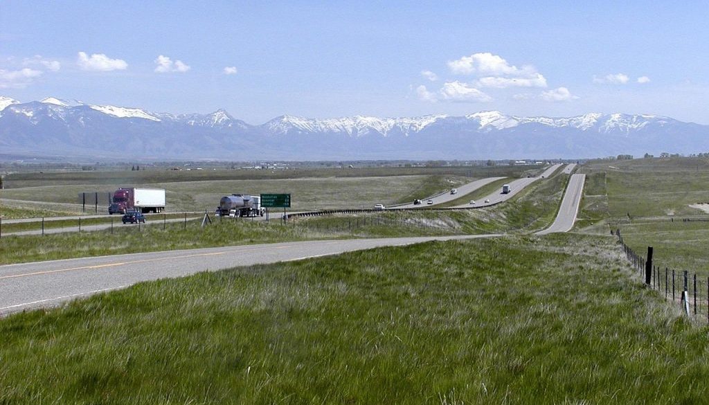 A long, open Montana highway framed by green fields and snow-capped mountains captures the pure freedom on the road that Montana vehicle registration makes possible.
