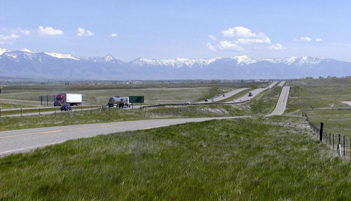 A long, open Montana highway framed by green fields and snow-capped mountains captures the pure freedom on the road that Montana vehicle registration makes possible.