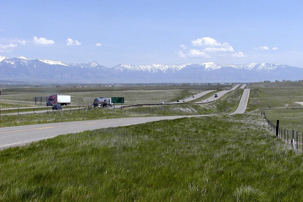 A long, open Montana highway framed by green fields and snow-capped mountains captures the pure freedom on the road that Montana vehicle registration makes possible.