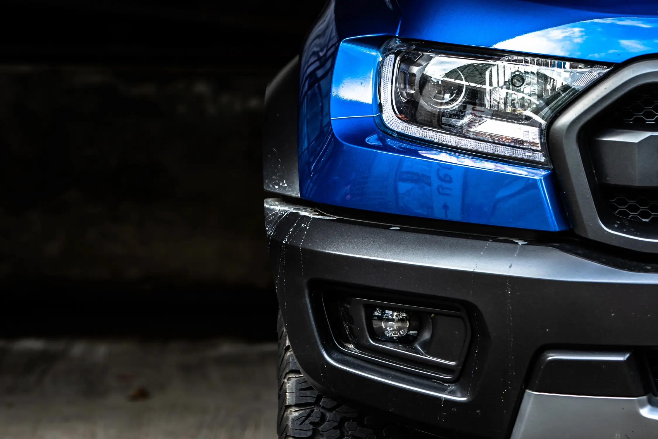 Close-up of a blue pickup truck front end showing headlight assembly, black grille, bumper, and fog light against a dark background.