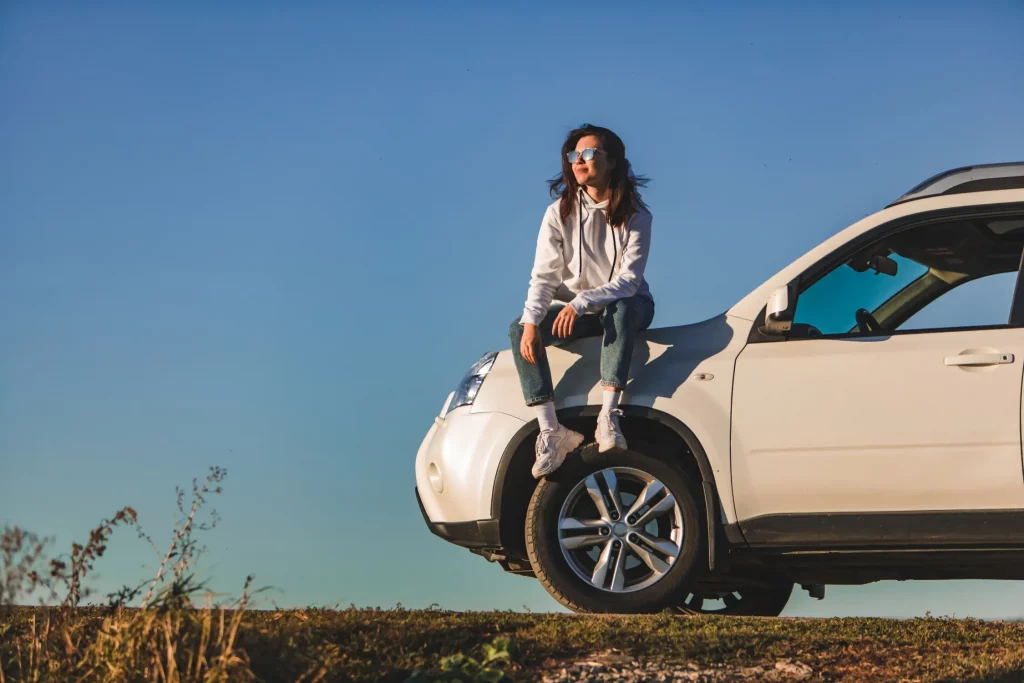 A happy woman sits on the front of her car, relaxed and smiling, enjoying total peace of mind after completing her LLC renewal.