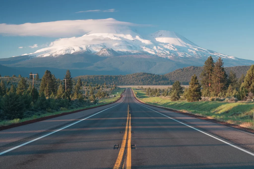 A straight two-lane highway leads toward a snow-covered mountain under a clear blue sky, reflecting the efficiency and reliability of Montana vehicle registration services for dealerships and commercial fleets.