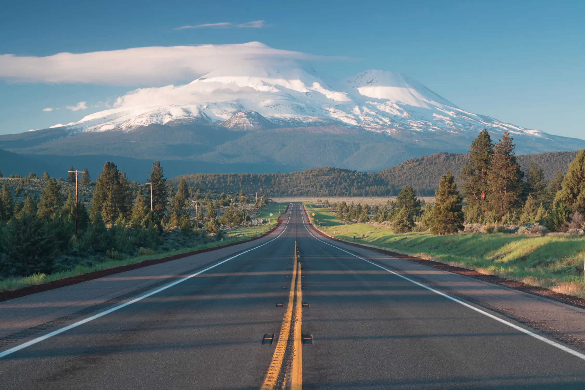 A straight two-lane highway leads toward a snow-covered mountain under a clear blue sky, reflecting the efficiency and reliability of Montana vehicle registration services for dealerships and commercial fleets.