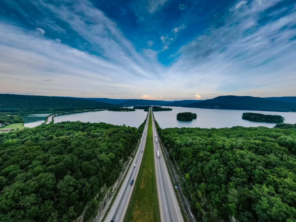 A straight highway slices through dense forest and a wide lake, leading toward distant hills under a dramatic, cloud-streaked sky.