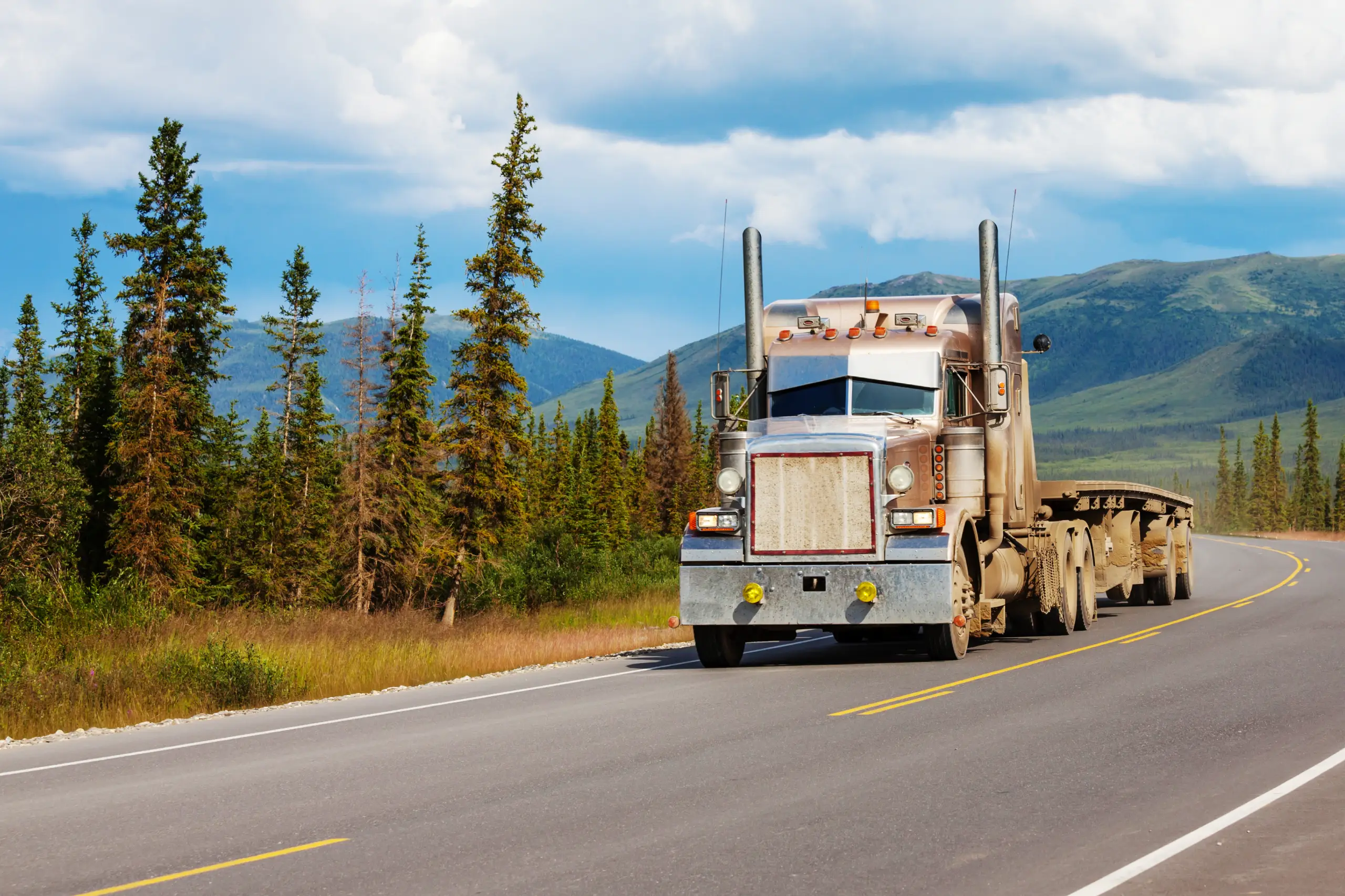 A semi-truck hauling a long flatbed trailer drives down a two-lane highway through a mountainous, forested landscape under a partly cloudy sky.