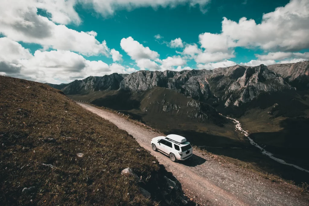 White SUV driving along a narrow mountain road overlooking a rugged alpine landscape with dramatic peaks and a partly cloudy sky.