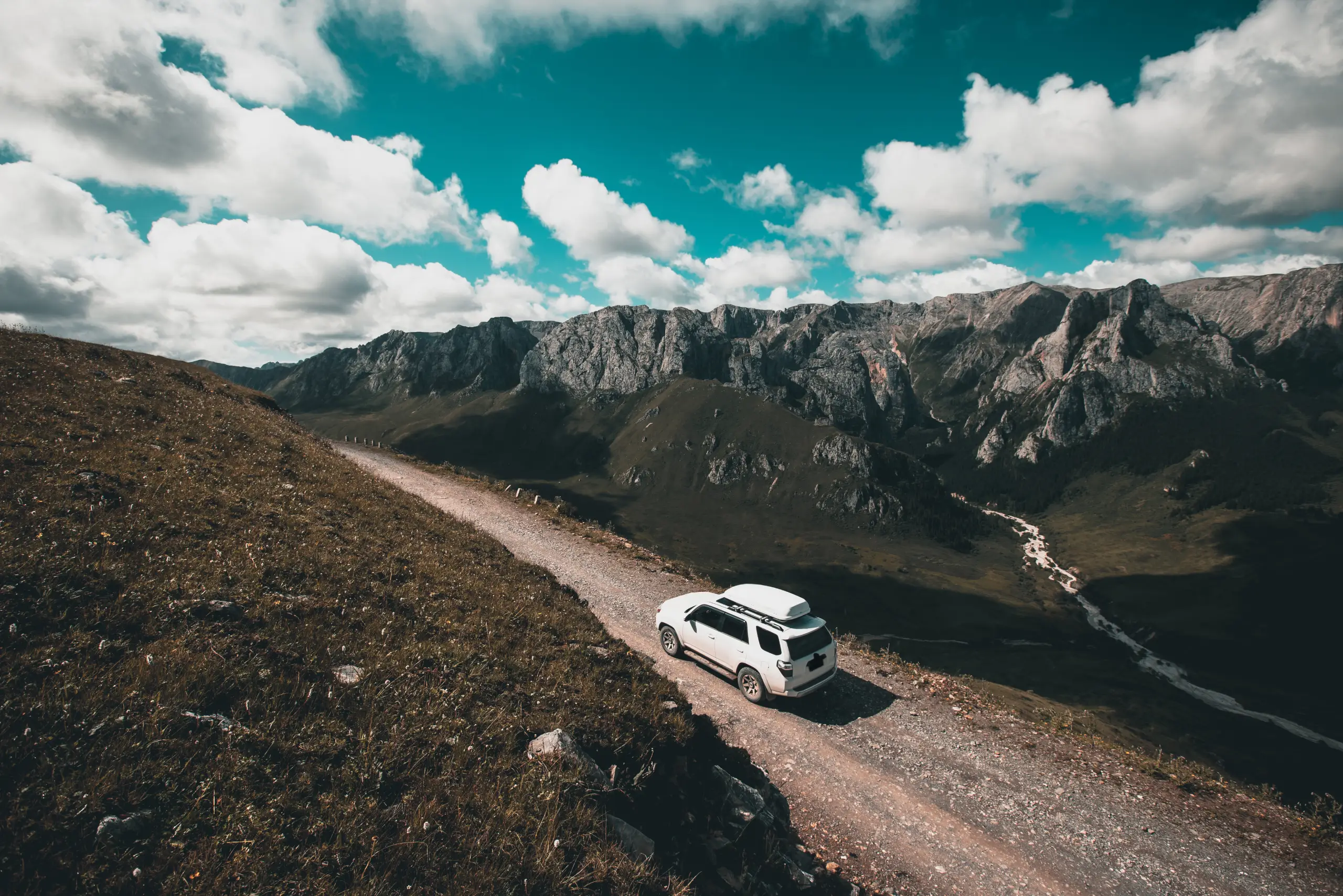 White SUV driving along a narrow mountain road overlooking a rugged alpine landscape with dramatic peaks and a partly cloudy sky.