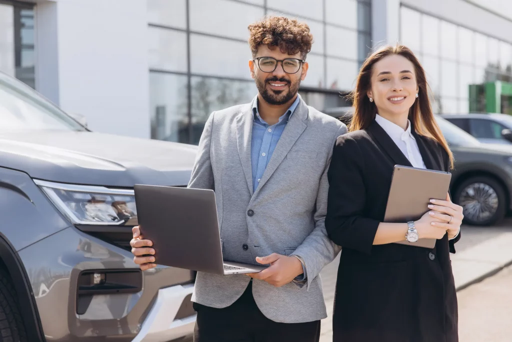 Two business professionals standing beside a vehicle outside a dealership, holding a laptop and tablet, represent structured and dependable Montana vehicle registration support for dealerships and fleet partners.