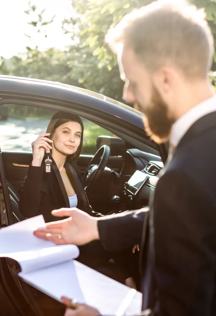 A driver sitting in her car hands documents and car keys to a professional outside the vehicle, illustrating an in-person vehicle handoff related to vehcile registration and paperwork processing.