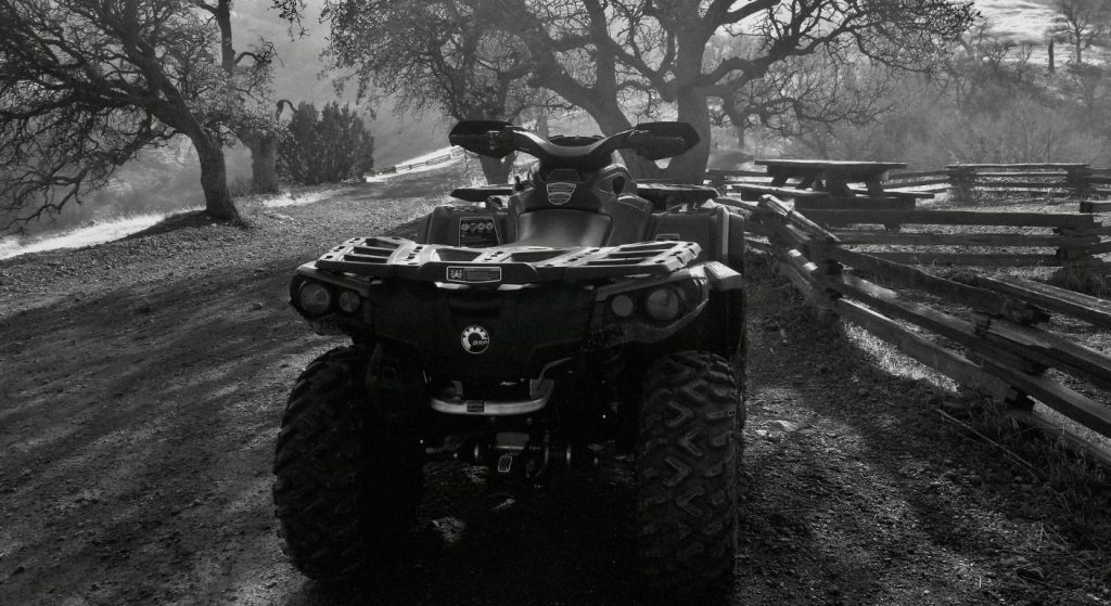 Front view of an ATV parked on a dirt road beside a wooden fence in a rural landscape