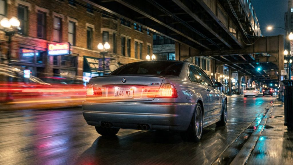 Silver BMW M3 driving on a wet city street at night, red taillights streaking with traffic blur.