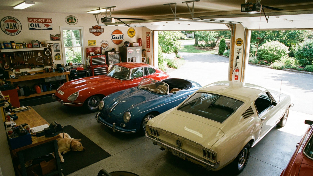 A high-angle interior view of a well-organized garage housing three classic cars: a red Jaguar E-Type, a blue Porsche 356 Speedster, and a cream-colored Ford Mustang Fastback. The garage features vintage automotive signage, a wooden workbench, and a yellow dog sleeping on the floor.