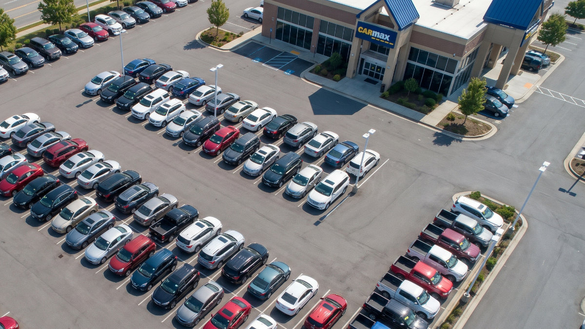 An aerial, high-angle shot of a CarMax dealership featuring rows of neatly parked colorful cars, SUVs, and trucks in a paved parking lot under a bright, sunny sky.