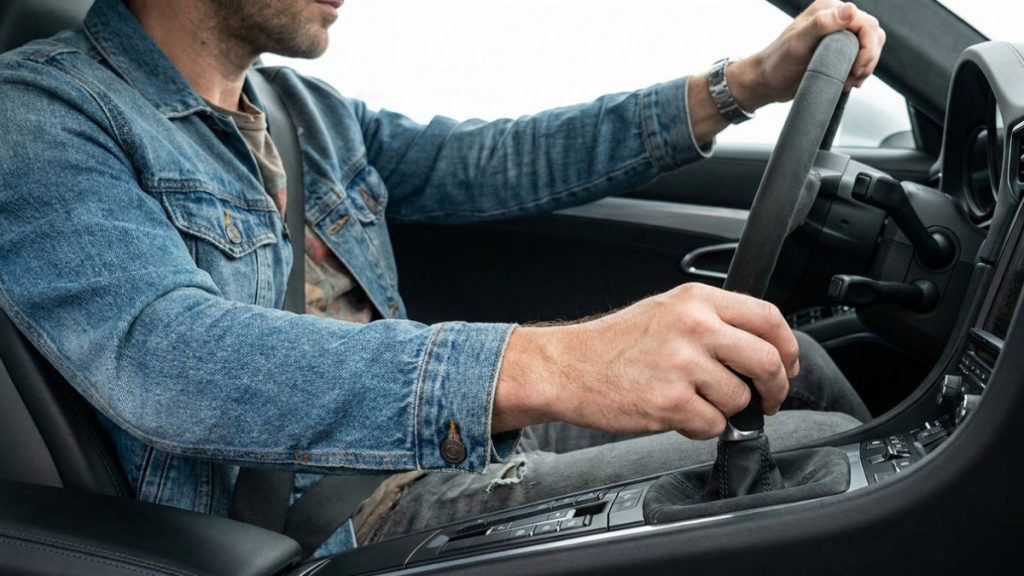 Interior close-up view focusing on a male driver's right hand actively gripping and moving a manual gear shift lever in a modern sports car. He wears a denim jacket.