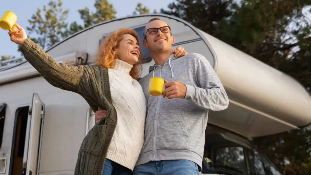 A happy couple stands in front of their RV holding coffee mugs, highlighting the freedom of RV ownership and the importance of proper motorhome registration and title.